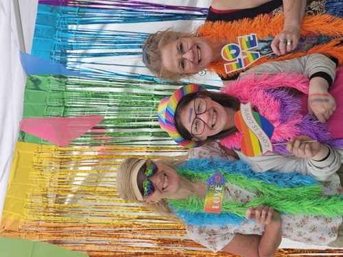 A photograph of three women across a range of ages wearing bright rainbow accessories against a rainbow background