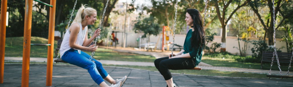 Two girls sat on a swing facing each other.