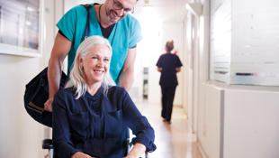 An elderly lady with grey hair being pushed in a wheelchair by a smiling man