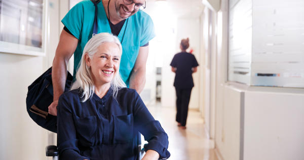 An elderly lady with grey hair being pushed in a wheelchair by a smiling man