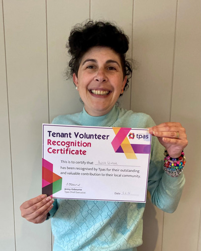 Photograph of Alisa, who has black hair with pale skin, wearing a blue jumper and holding up with Tenant Volunteer Recognition Certificate, awarded to her by Tpas.