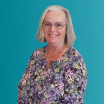 Mary, Customer Feedback Panel member, wearing a flowery top, glasses against a teal coloured backdrop.